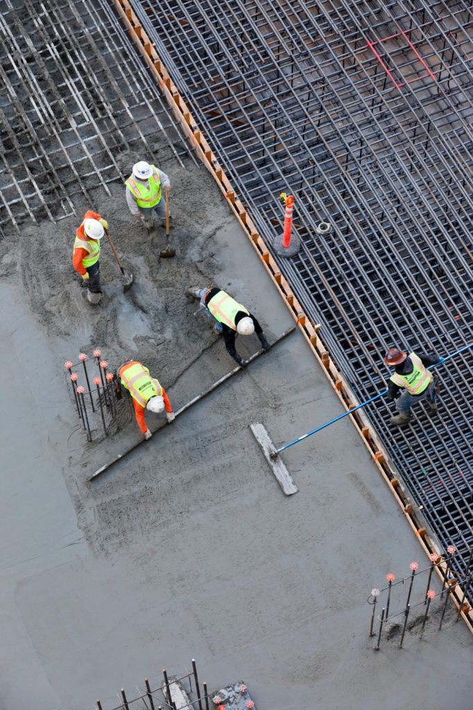 An overhead shot shows five construction workers laying a concrete foundation