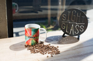A mug of coffee with the SFMOMA logo, next to a pile of coffee beans and a sign reading "Sight Glass"