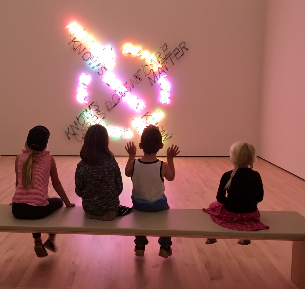 Four children sit on a bench in front of Bruce Nauman's Life Death/Knows Doesn't Know